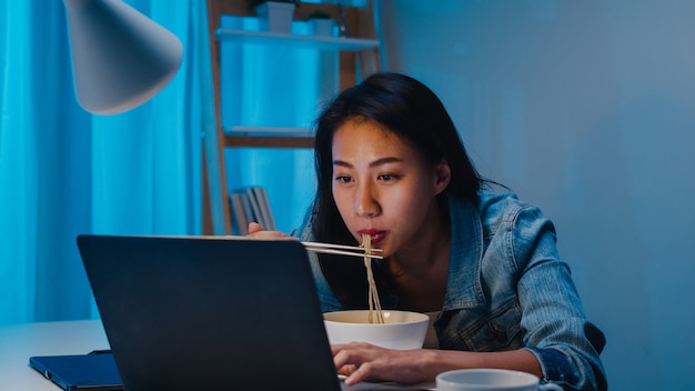 Free photo asia freelance smart business women eating instant noodles while working on laptop in living room at home at night. happy young asian girl sitting on desk work overtime, enjoy relax time.